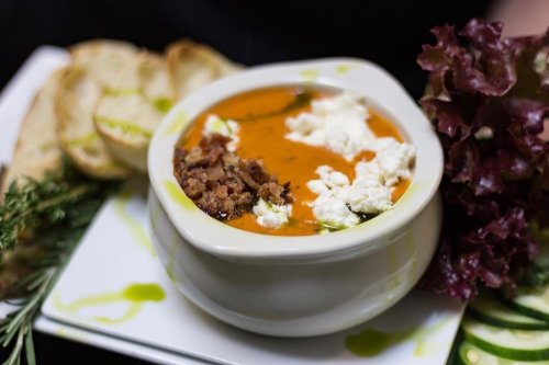 A bowl of creamy soup topped with crumbled bacon and cheese, served with bread and fresh greens on a plate.