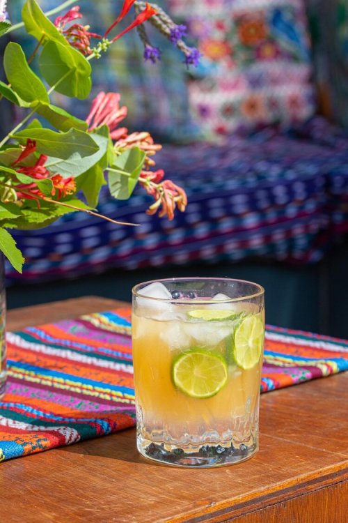 A refreshing cocktail with lime slices sits on a wooden table, surrounded by colorful textiles and a floral arrangement.