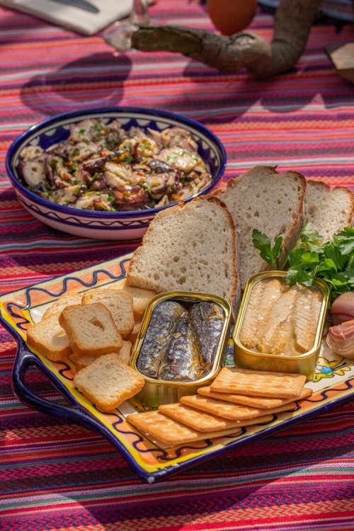 A colorful platter with bread, crackers, canned fish, and a bowl of salad on a vibrant tablecloth.