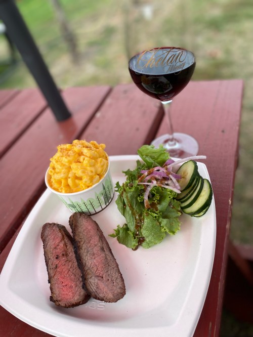A plate with sliced beef, macaroni and cheese, salad with cucumbers, and a glass of red wine on a wooden table.