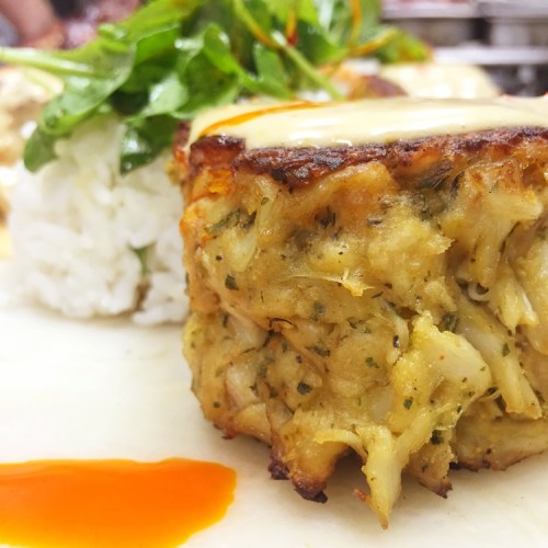 A close-up of a crab cake topped with melted cheese, served with rice and garnished with fresh greens.