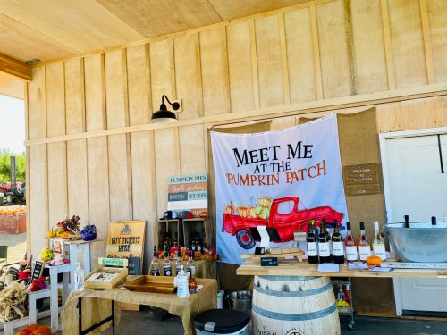 A rustic pumpkin patch stand with a banner, pumpkins, wine bottles, and a wooden table in a cozy setting.