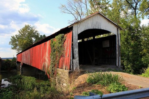 A weathered red covered bridge surrounded by trees and greenery, with a gravel path leading to it.