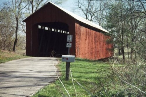 A red covered bridge stands beside a gravel road, with a mailbox nearby and trees in the background.