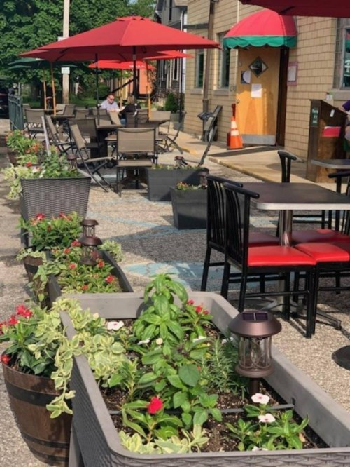 Outdoor dining area with red umbrellas, planters, and tables along a street, creating a cozy atmosphere.