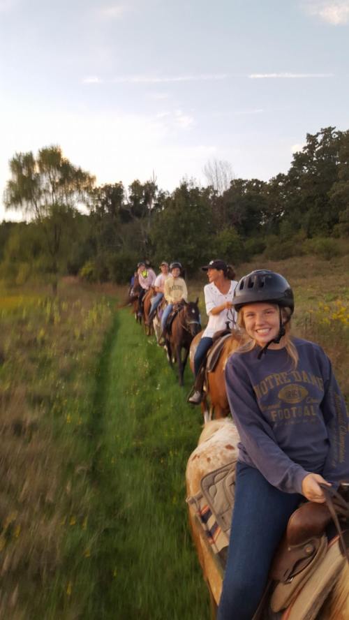 A group of people riding horses along a grassy trail during sunset, with one person smiling in the foreground.
