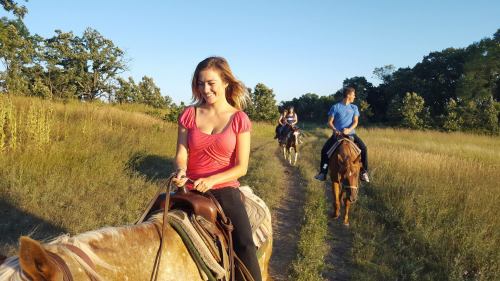 Two people riding horses on a grassy trail, with a woman in a pink shirt smiling in the foreground.