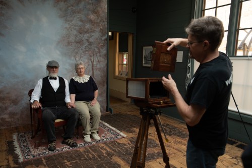 A photographer prepares to take a portrait of a couple seated in a vintage studio with a backdrop.
