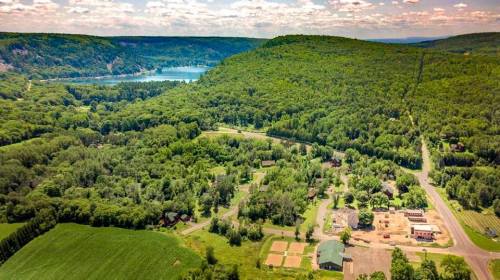 Aerial view of lush green hills, a river, and a small town surrounded by trees and fields under a partly cloudy sky.