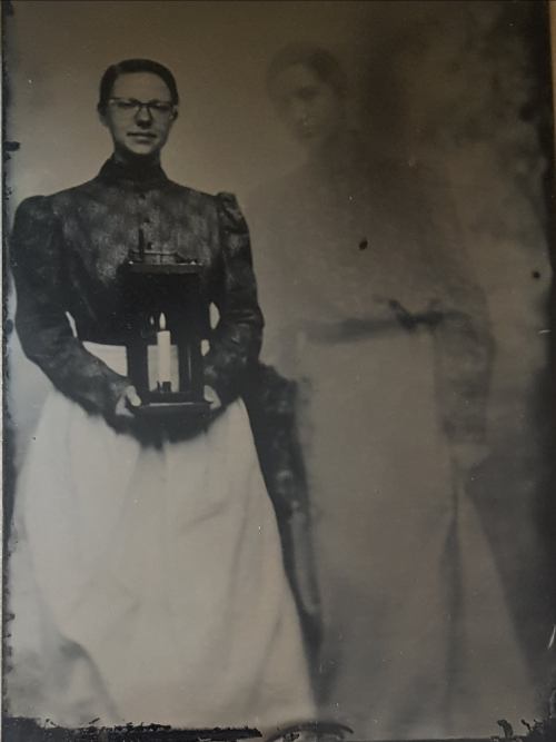 A vintage portrait of a woman in period clothing holding a lantern, with a ghostly figure in the background.