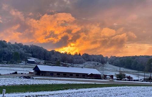 A snowy landscape at sunset, with vibrant orange clouds above a farm building and rolling hills in the background.
