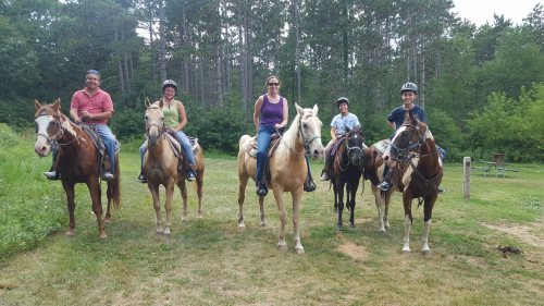 A group of five people on horseback, smiling, in a green outdoor setting with trees in the background.