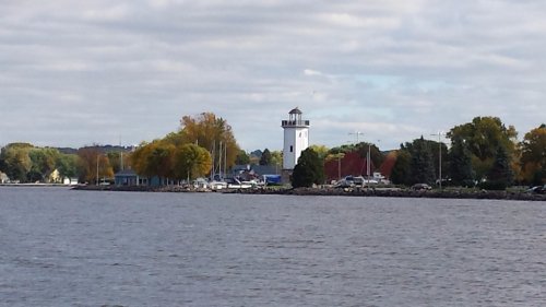A scenic view of a waterfront with a lighthouse, colorful trees, and boats along the shore under a cloudy sky.