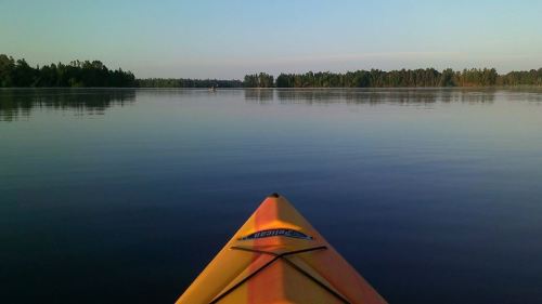 A kayak's bow on calm water, reflecting trees and a clear sky in the distance.