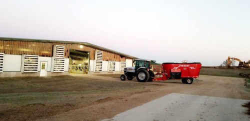 A tractor with a red feed mixer parked outside a barn at dusk, with a clear sky and gravel driveway.