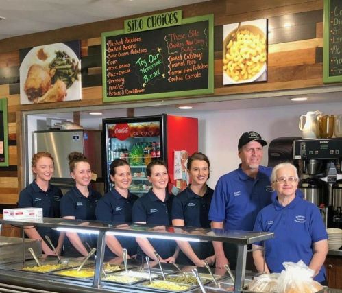 A group of seven smiling staff members stands behind a food counter in a casual dining setting.