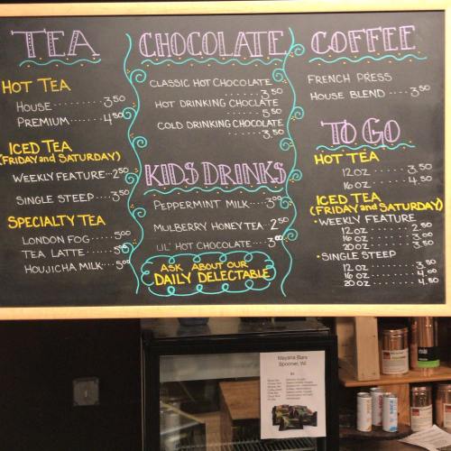 Chalkboard menu featuring tea, chocolate, coffee, and kids' drinks with prices and weekly specials listed.