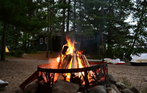 A campfire burns brightly in a forested area, with a cabin and boats visible in the background.