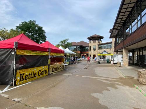 A market scene with colorful tents, including a kettle corn stand, and people walking near a building.