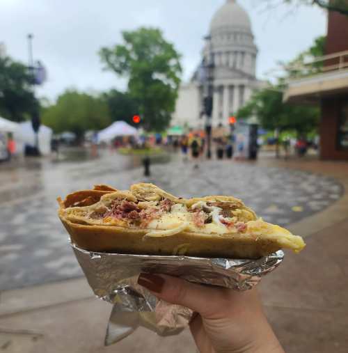 A hand holds a wrapped burrito in front of a rainy outdoor event with a large building in the background.