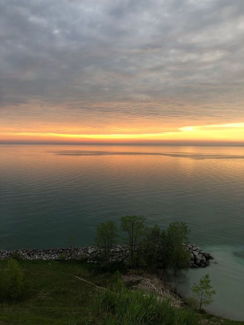 A serene lake at sunset, with calm waters reflecting orange and gray skies, framed by greenery and rocky shoreline.