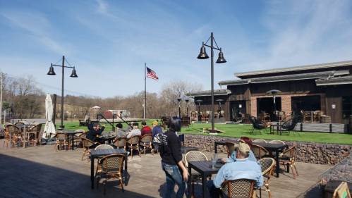 Outdoor seating area of a restaurant with people dining, an American flag, and a clear blue sky in the background.