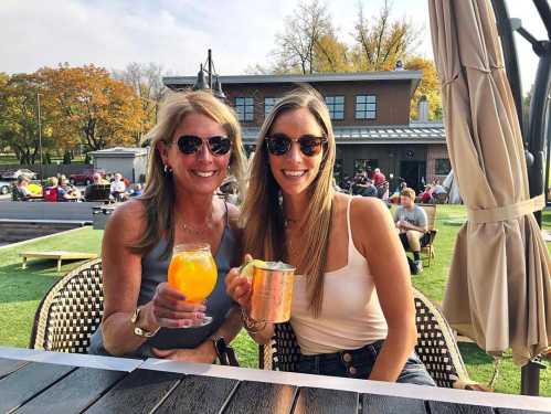 Two women in sunglasses smile while holding drinks at an outdoor setting with greenery and people in the background.