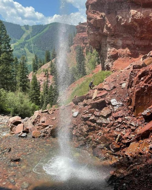 A waterfall cascades down rocky terrain surrounded by lush green trees and mountains under a clear blue sky.