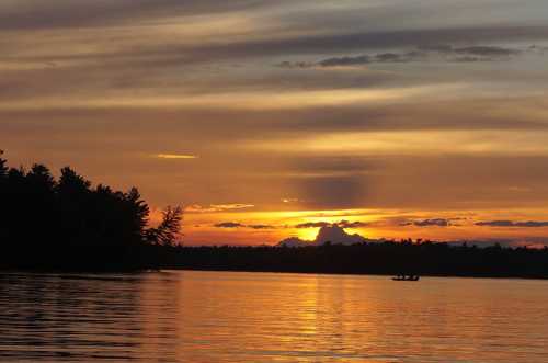 A serene sunset over a lake, with vibrant orange and purple hues reflecting on the water and silhouetted trees in the foreground.