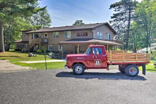 A vintage red truck parked in front of a two-story brown building surrounded by trees and a grassy area.