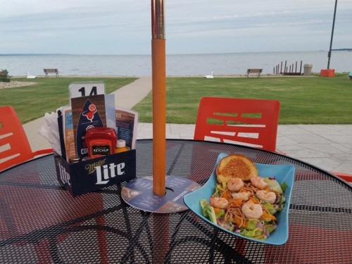A table by the water featuring a salad with shrimp, condiments, and a scenic view of the lake.