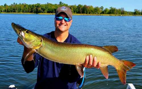 A smiling person holds a large fish while standing on a boat in a lake surrounded by trees.