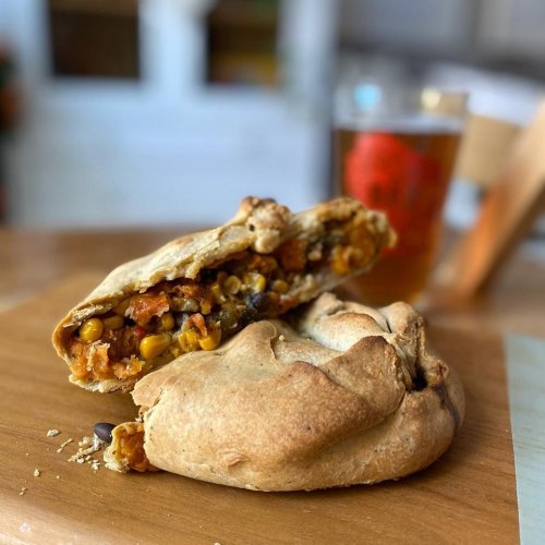 A close-up of a flaky pastry filled with vegetables and grains, resting on a wooden surface next to a glass of beer.