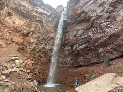 A tall waterfall cascades down rocky cliffs, with a person standing near the base in a scenic, rugged landscape.