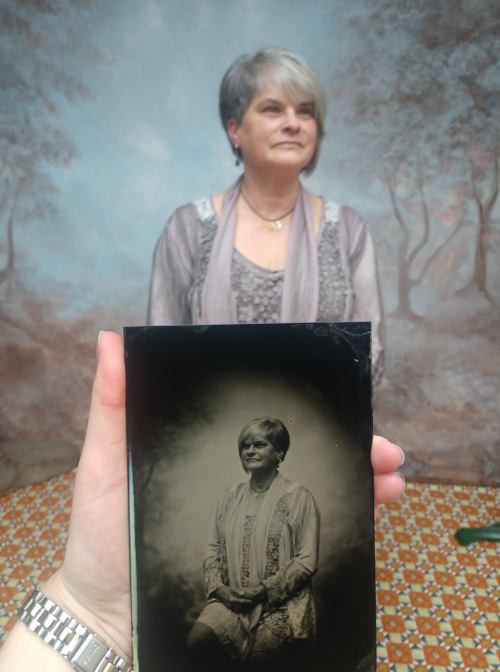 A person holds a vintage photo of a woman, while the woman stands in the background, both in a scenic setting.
