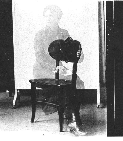 A ghostly figure of a woman seated on a chair, holding a piece of paper, with a blurred background.