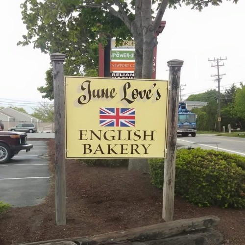 Sign for "June Love's English Bakery" featuring a British flag, with a tree and parked car in the background.