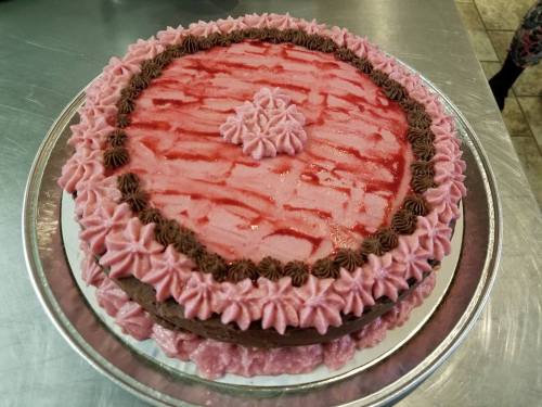 A round cake with pink frosting, chocolate accents, and a decorative design on top, placed on a silver platter.