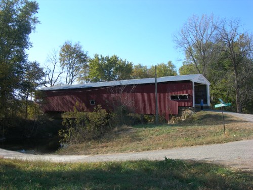 A red covered bridge stands beside a gravel road, surrounded by trees and greenery under a clear blue sky.