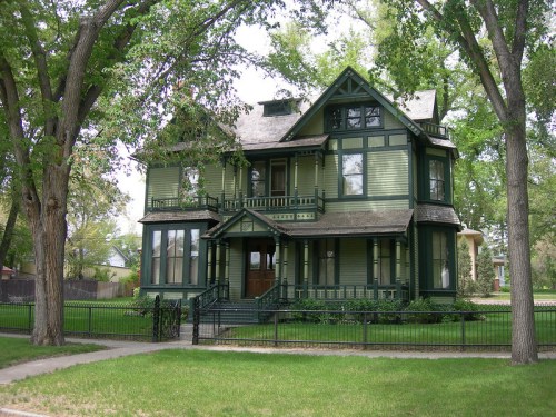 A large, green Victorian-style house surrounded by trees and a grassy lawn, featuring intricate architectural details.