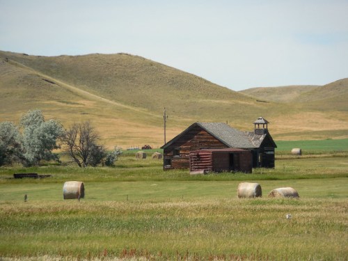 A rustic wooden barn surrounded by hay bales and rolling green hills under a clear sky.