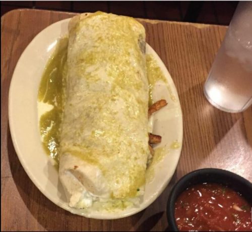 A large burrito topped with green sauce, served on a plate with a side of salsa and a glass of water.