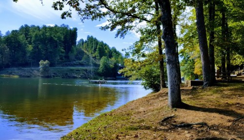 A serene lake surrounded by trees, with a dock in the distance and a clear blue sky reflecting on the water.
