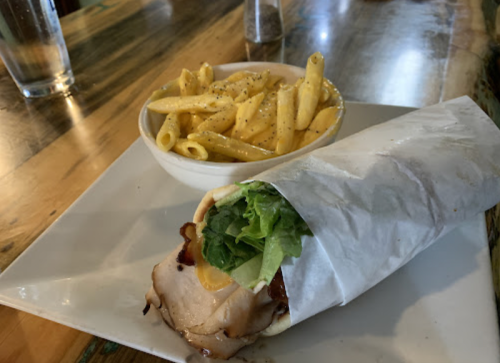 A wrapped sandwich with turkey and lettuce beside a bowl of pasta with seasoning on a wooden table.