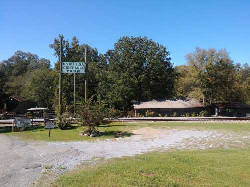 Sign for Kymulga Grist Mill Park with trees and buildings in the background on a clear day. Gravel path in the foreground.