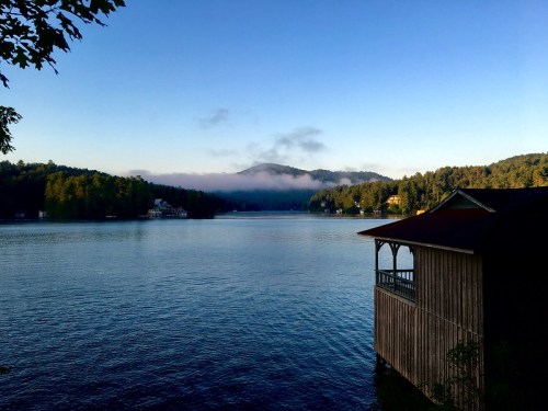 A serene lake view at dawn, with mist rising over the water and a wooden cabin on the shore surrounded by trees.