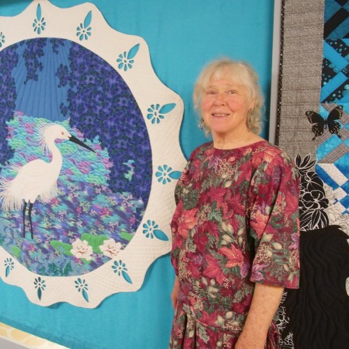 A smiling woman stands beside a colorful quilt featuring a white heron and floral designs.