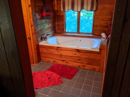 Cozy wooden bathroom featuring a large tub, window with a view, and red towels on the floor.