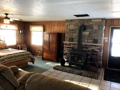 Cozy cabin interior featuring a bed, stone fireplace, wooden furniture, and natural light from windows.