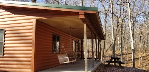 A wooden cabin with a porch swing, surrounded by trees in a wooded area.